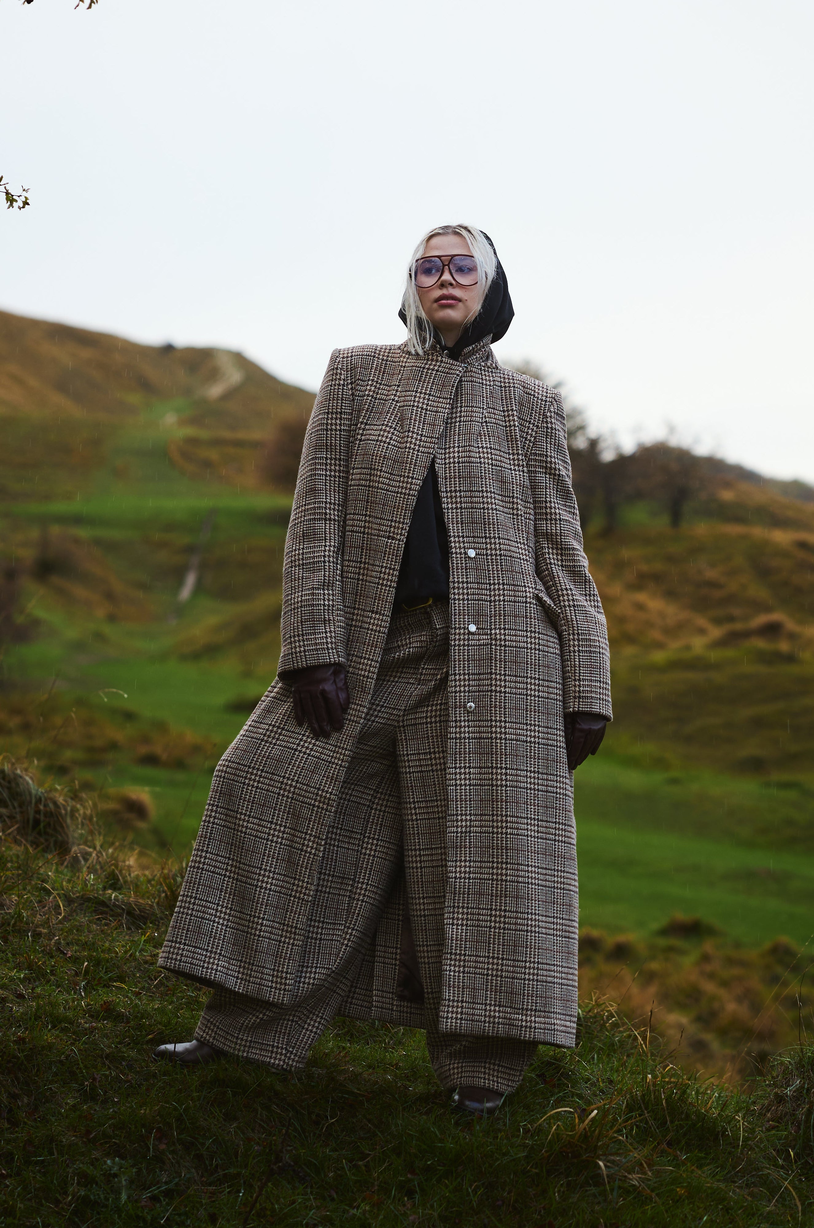 Person wearing a long coat standing in a grassy field with hills in the background